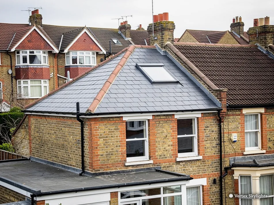 A hipped slate roof with a roof window above a yellow-brick house and flat-roofed extension