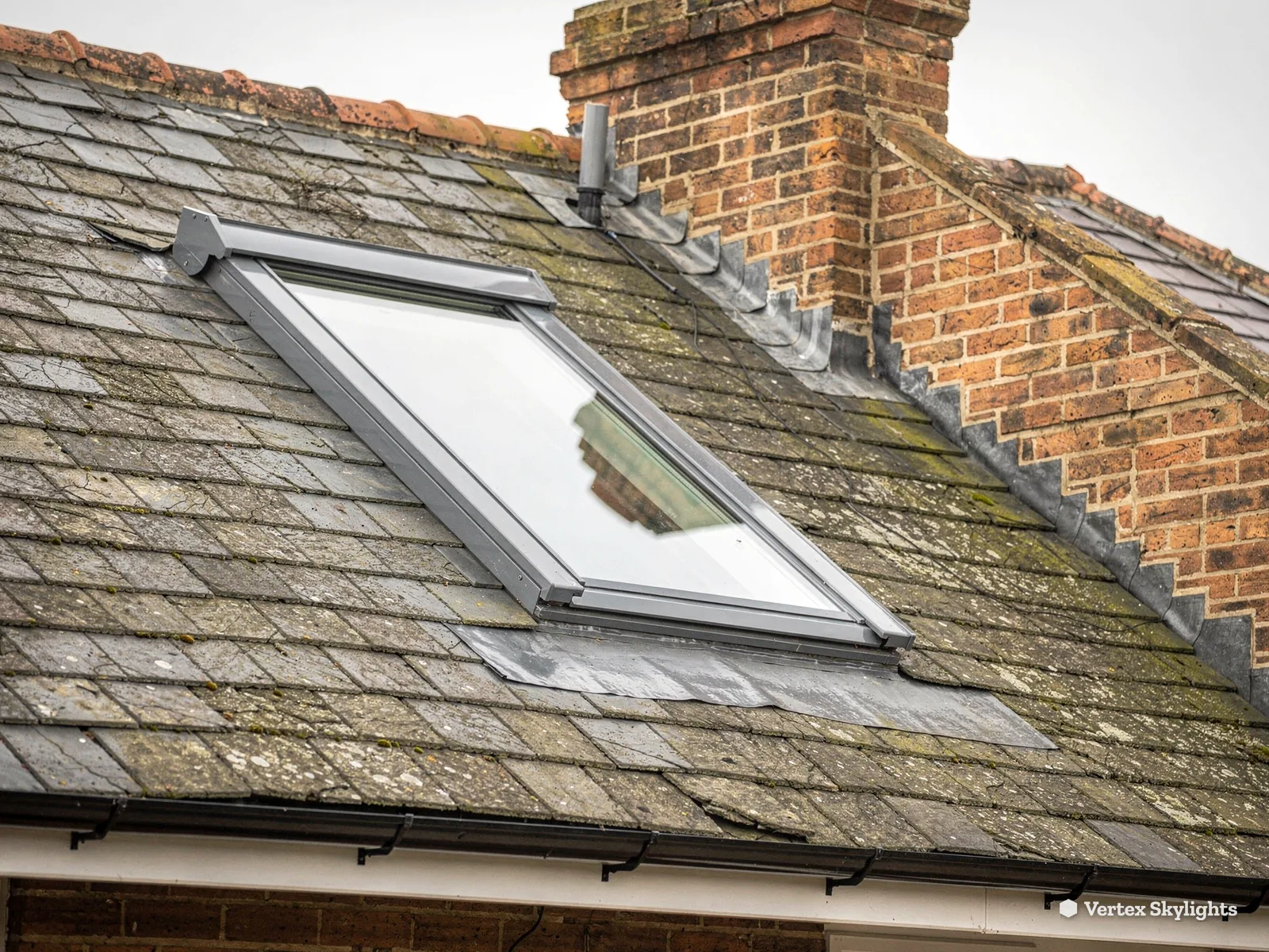 A roof window in a weathered slate roof beside a brick chimney